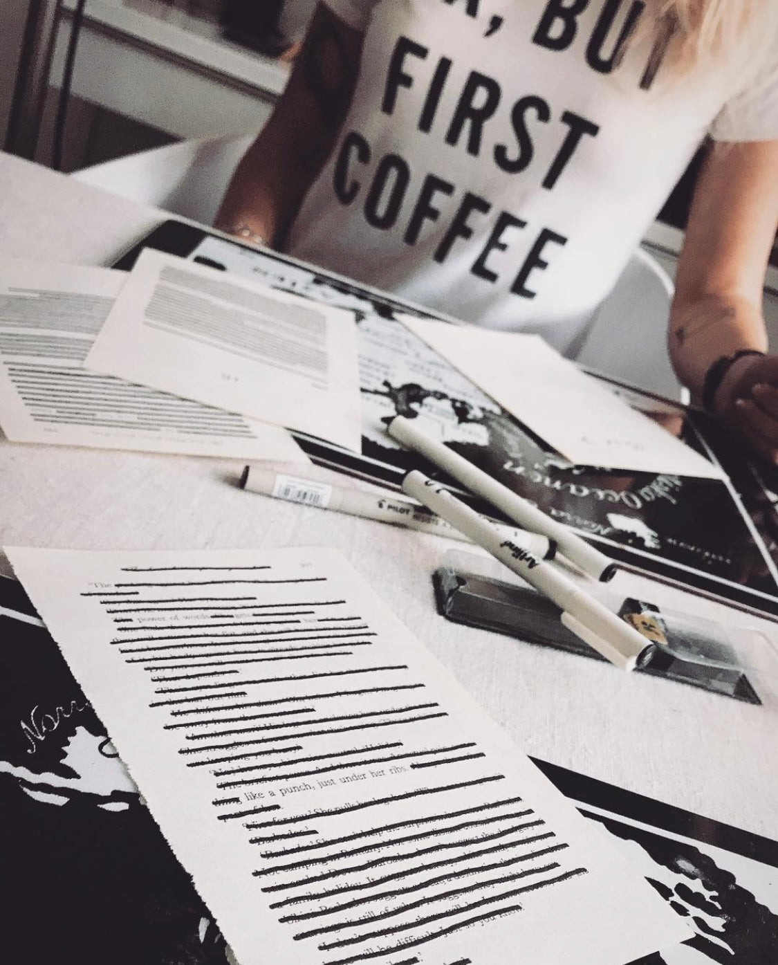 Person wearing a 'Ok, But First Coffee' shirt with a desk setup featuring papers and pens, creating blackout poetry.